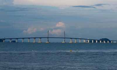 Long bridge spanning a calm sea with a central cable-stayed section and a small island in the distance.