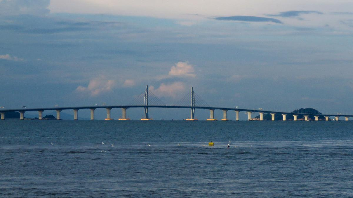 Long bridge spanning a calm sea with a central cable-stayed section and a small island in the distance.