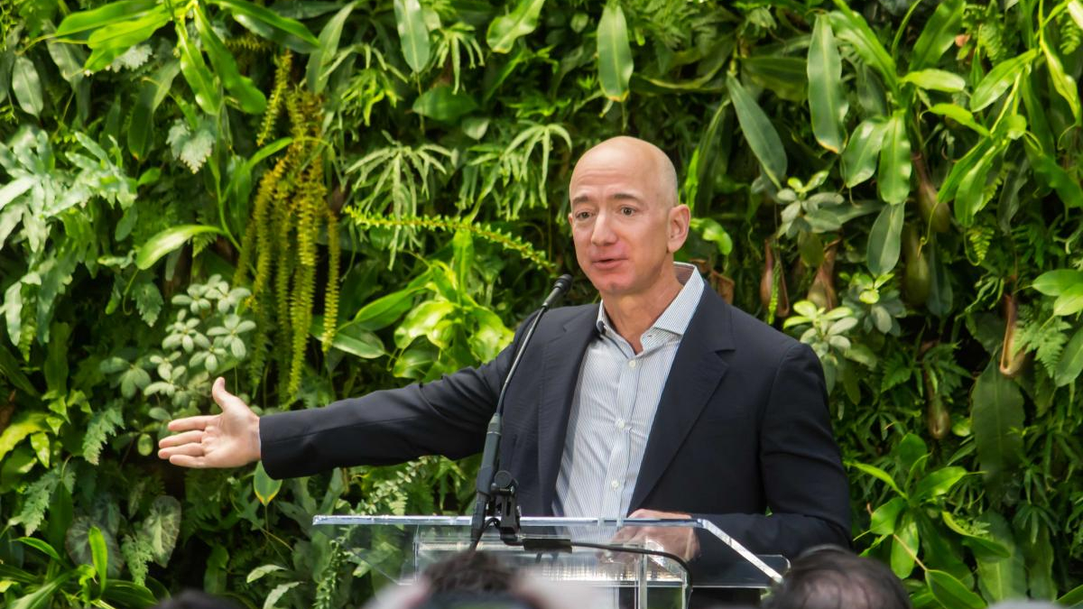 Man in a dark suit and light shirt speaks at a clear podium with a microphone, gesturing with his left arm, lush green plants behind him.