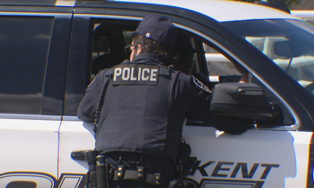 Back view of a police officer in a vest labeled POLICE leaning into a white-and-black patrol SUV parked at the curb.