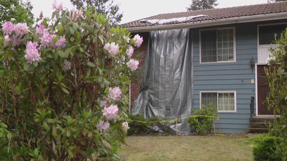Blue house with a large gray tarp covering a section of the wall, flanked by pink rhododendron flowers in the foreground and yellow caution tape on the ground.