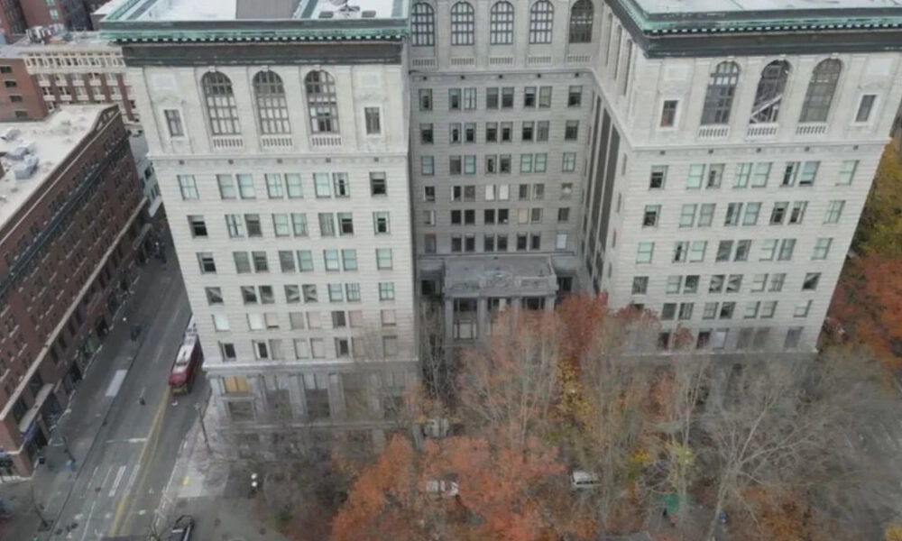 Aerial view of a large pale stone building with a central entrance, surrounded by a courtyard and autumn-colored trees; city streets and other buildings visible nearby.