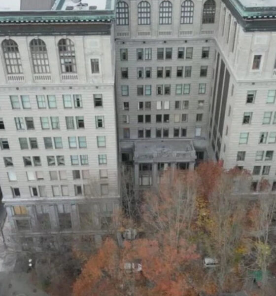Aerial view of a large pale stone building with a central entrance, surrounded by a courtyard and autumn-colored trees; city streets and other buildings visible nearby.