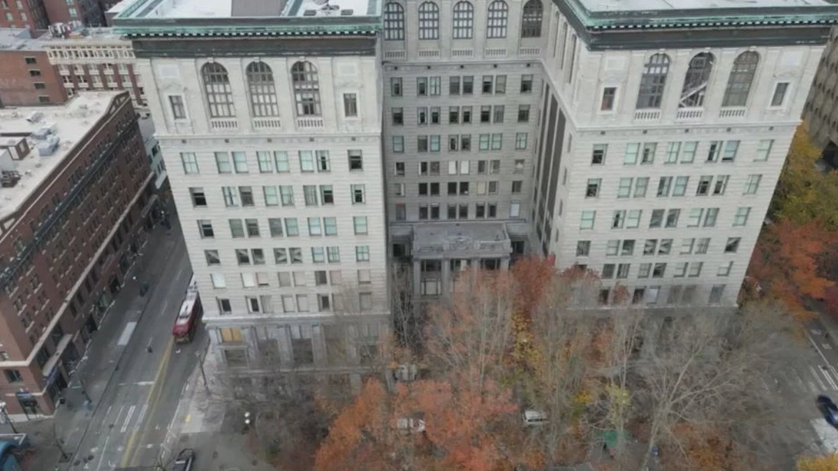 Aerial view of a large pale stone building with a central entrance, surrounded by a courtyard and autumn-colored trees; city streets and other buildings visible nearby.