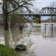 Flooded riverside with a metal truss bridge, bare trees, and reflective water surface.