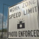 Large road work zone sign stating 'Work Zone Speed Limit' with 'Photo Enforced' at a construction site under a blue sky.