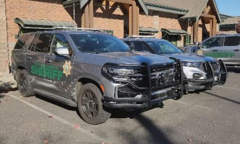 Two gray sheriff patrol SUVs with heavy front bumpers parked in a lot beside a brick building.