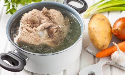 Pot of simmering beef soup with chunks of meat, surrounded by potato, carrot, onion, and garlic on a white cutting board.