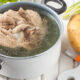Pot of simmering beef soup with chunks of meat, surrounded by potato, carrot, onion, and garlic on a white cutting board.