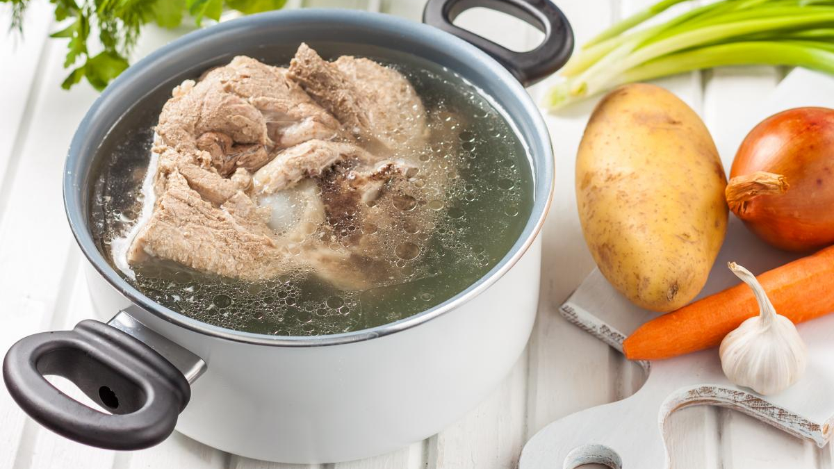 Pot of simmering beef soup with chunks of meat, surrounded by potato, carrot, onion, and garlic on a white cutting board.