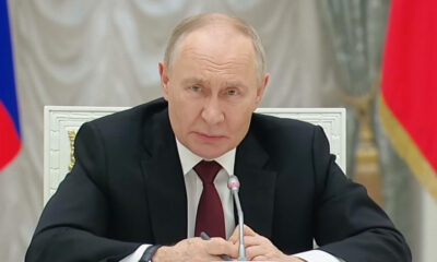 A man in a dark suit and red tie sits at a desk with a microphone in front, speaking seriously at a formal meeting.