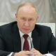 A man in a dark suit and red tie sits at a desk with a microphone in front, speaking seriously at a formal meeting.
