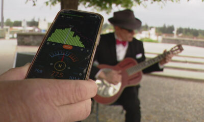 Person holds a smartphone showing an audio app with meters while a guitarist in a hat plays a pink guitar in the background.