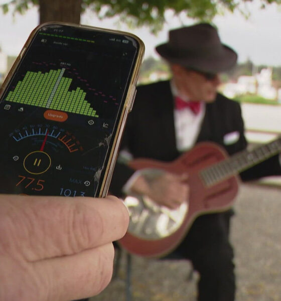 Person holds a smartphone showing an audio app with meters while a guitarist in a hat plays a pink guitar in the background.