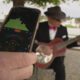 Person holds a smartphone showing an audio app with meters while a guitarist in a hat plays a pink guitar in the background.