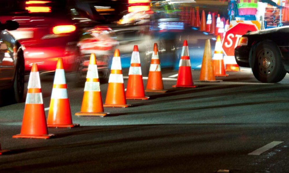 Nighttime roadwork scene with orange traffic cones lining a construction zone and blurred cars in motion as they pass by.