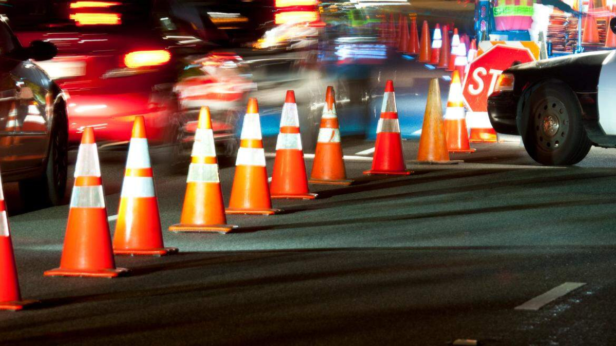 Nighttime roadwork scene with orange traffic cones lining a construction zone and blurred cars in motion as they pass by.