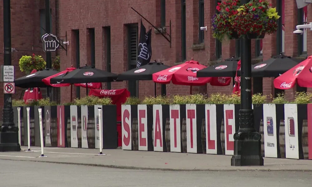 Sidewalk cafe scene with red Coca-Cola umbrellas and a row of white panels spelling SEATTLE along the railing, brick buildings in the background, and hanging flower baskets above.