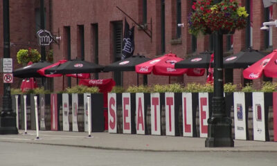 Sidewalk cafe scene with red Coca-Cola umbrellas and a row of white panels spelling SEATTLE along the railing, brick buildings in the background, and hanging flower baskets above.