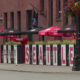 Sidewalk cafe scene with red Coca-Cola umbrellas and a row of white panels spelling SEATTLE along the railing, brick buildings in the background, and hanging flower baskets above.