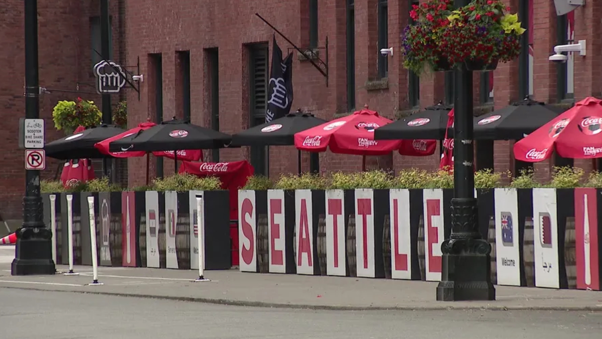 Sidewalk cafe scene with red Coca-Cola umbrellas and a row of white panels spelling SEATTLE along the railing, brick buildings in the background, and hanging flower baskets above.
