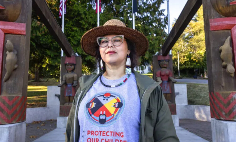 Person wearing a straw hat and glasses stands in a park with carved totem-like structures in the background, wearing a gray shirt that reads 'Protecting Our Children'.