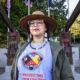 Person wearing a straw hat and glasses stands in a park with carved totem-like structures in the background, wearing a gray shirt that reads 'Protecting Our Children'.