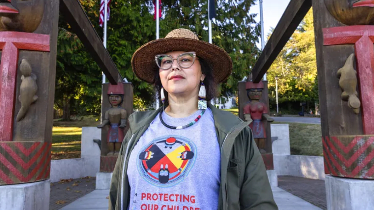 Person wearing a straw hat and glasses stands in a park with carved totem-like structures in the background, wearing a gray shirt that reads 'Protecting Our Children'.