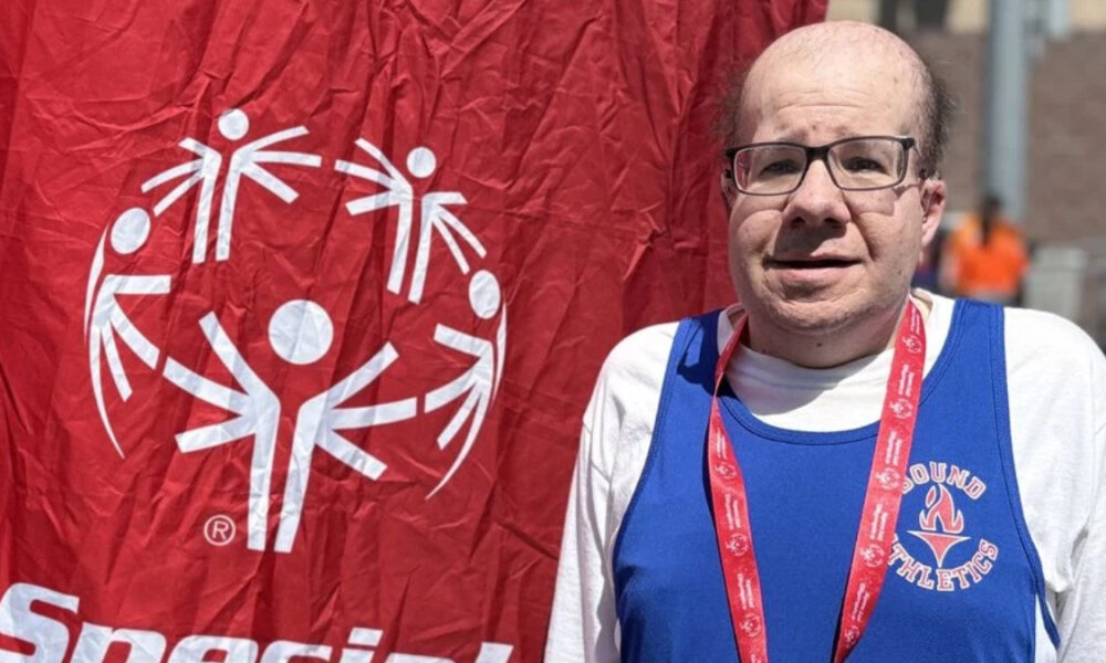 Man with glasses in a blue athletic jersey and red lanyard posing beside a red banner with a white circular logo emblem behind him.
