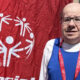 Man with glasses in a blue athletic jersey and red lanyard posing beside a red banner with a white circular logo emblem behind him.