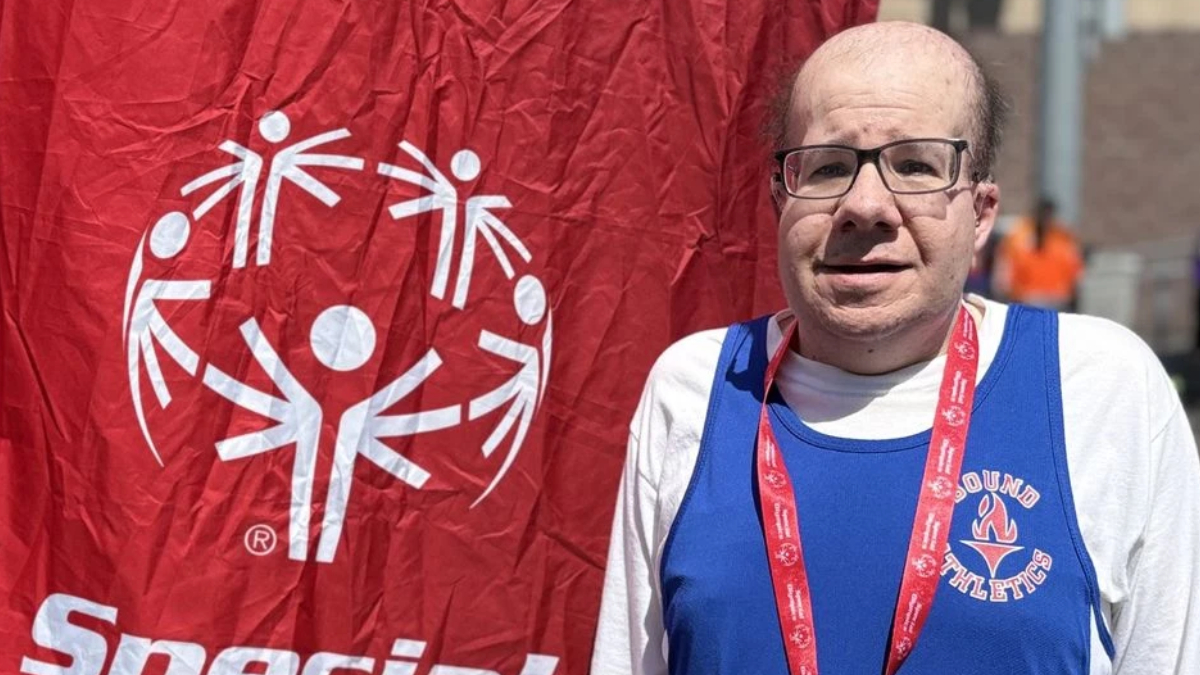 Man with glasses in a blue athletic jersey and red lanyard posing beside a red banner with a white circular logo emblem behind him.