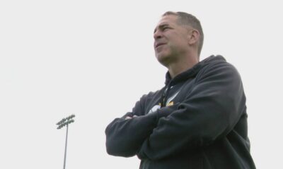 Man in a black hoodie stands with arms crossed on a sports field, looking off into the distance.