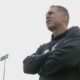 Man in a black hoodie stands with arms crossed on a sports field, looking off into the distance.