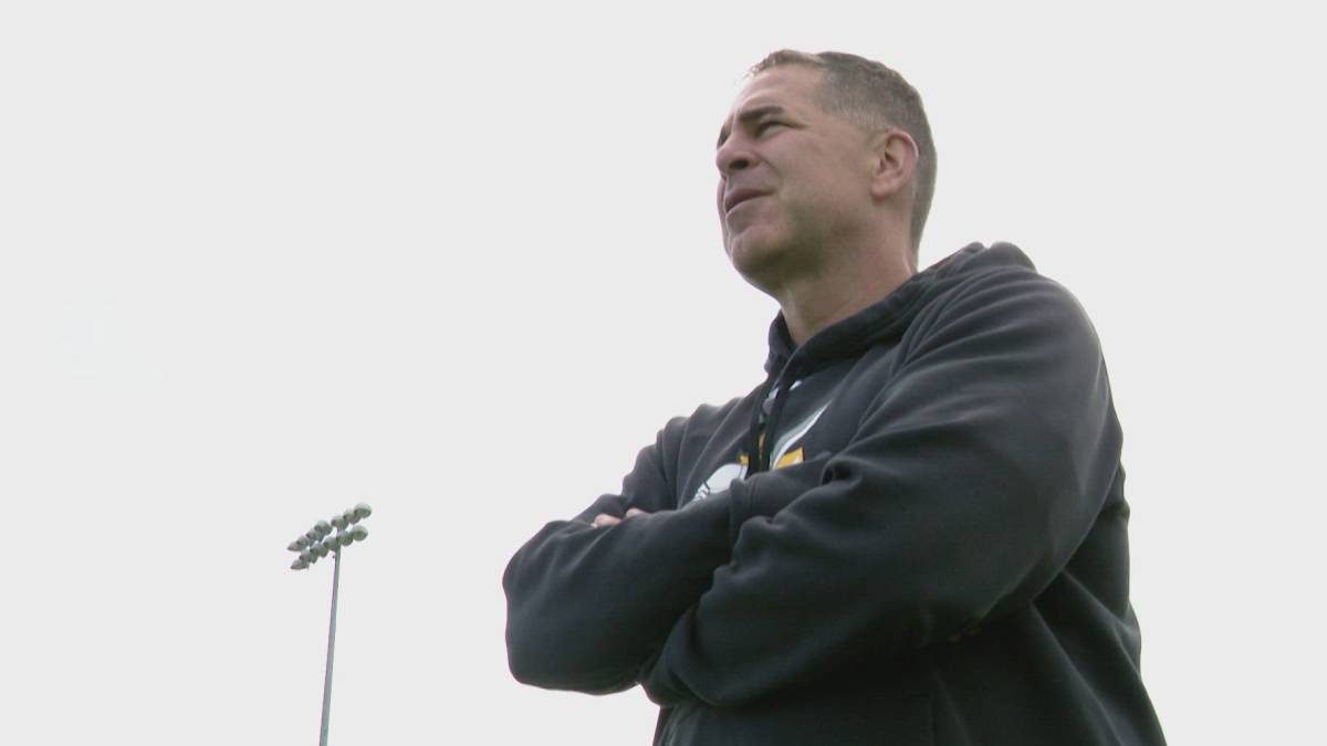 Man in a black hoodie stands with arms crossed on a sports field, looking off into the distance.