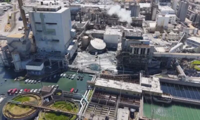 Aerial view of a large industrial complex with smokestacks, pipelines, and concrete processing buildings beside a water-filled area and parked cars nearby