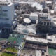 Aerial view of a large industrial complex with smokestacks, pipelines, and concrete processing buildings beside a water-filled area and parked cars nearby