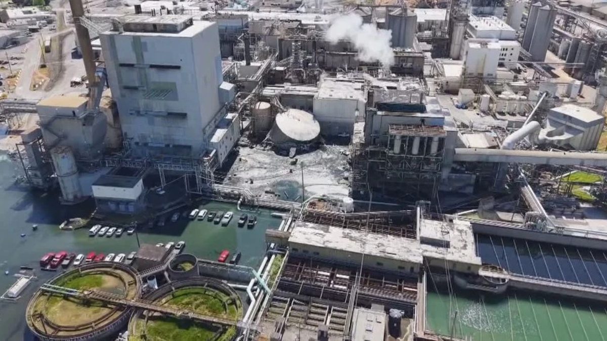 Aerial view of a large industrial complex with smokestacks, pipelines, and concrete processing buildings beside a water-filled area and parked cars nearby