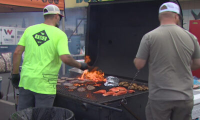 Two men grill burgers and salmon on a large outdoor barbecue, flames visible.