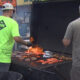 Two men grill burgers and salmon on a large outdoor barbecue, flames visible.