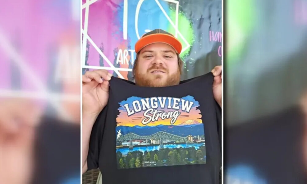 Man with a red cap holding up a black T-shirt that says ‘Longview Strong’ with a colorful landscape graphic in front of a mural backdrop.