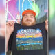 Man with a red cap holding up a black T-shirt that says ‘Longview Strong’ with a colorful landscape graphic in front of a mural backdrop.
