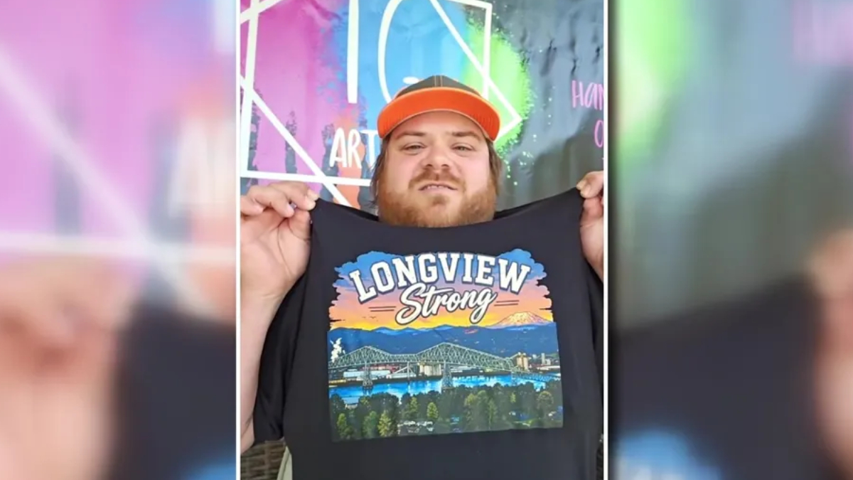 Man with a red cap holding up a black T-shirt that says ‘Longview Strong’ with a colorful landscape graphic in front of a mural backdrop.