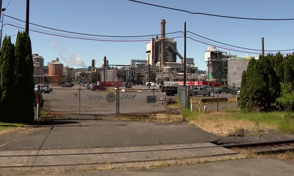 Industrial plant behind a chain-link fence with a gate, cars, and utility lines under a blue sky; railroad tracks in the foreground.