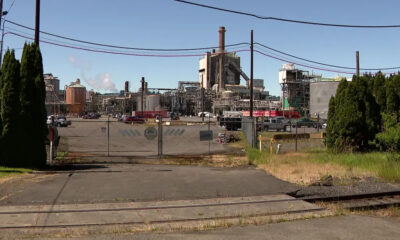 Industrial plant behind a chain-link fence with a gate, cars, and utility lines under a blue sky; railroad tracks in the foreground.