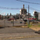 Industrial plant behind a chain-link fence with a gate, cars, and utility lines under a blue sky; railroad tracks in the foreground.