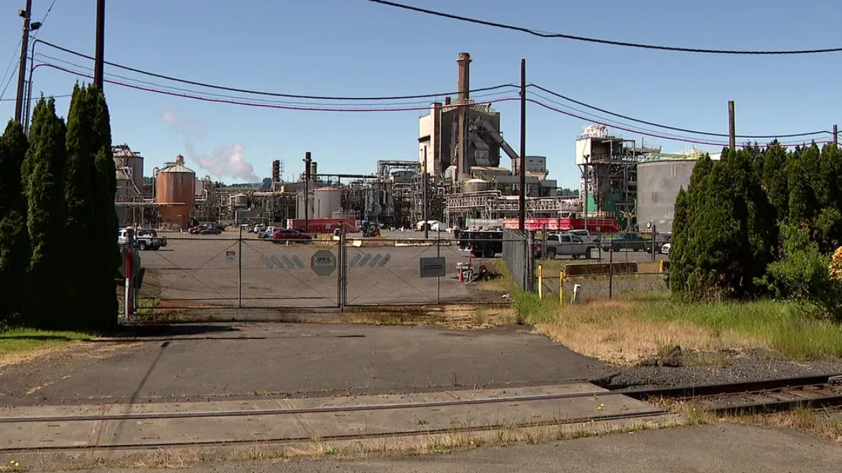 Industrial plant behind a chain-link fence with a gate, cars, and utility lines under a blue sky; railroad tracks in the foreground.