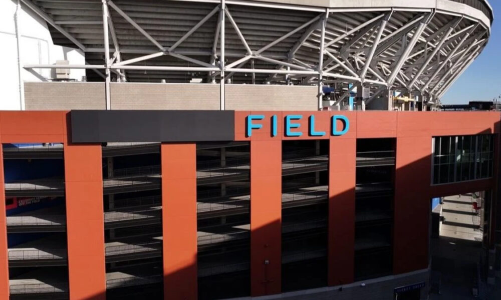 Exterior view of a stadium with an orange facade, multi-level parking, and a blue SIGN reading FIELD on top of the building.