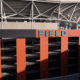 Exterior view of a stadium with an orange facade, multi-level parking, and a blue SIGN reading FIELD on top of the building.