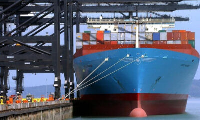 Large blue container ship docked at a port with stacked colorful containers and overhead gantry cranes.
