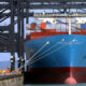 Large blue container ship docked at a port with stacked colorful containers and overhead gantry cranes.